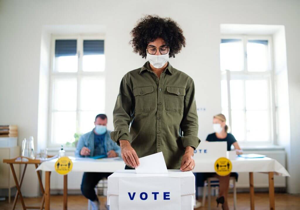 Young man putting his vote in the ballot box, elections and coronavirus