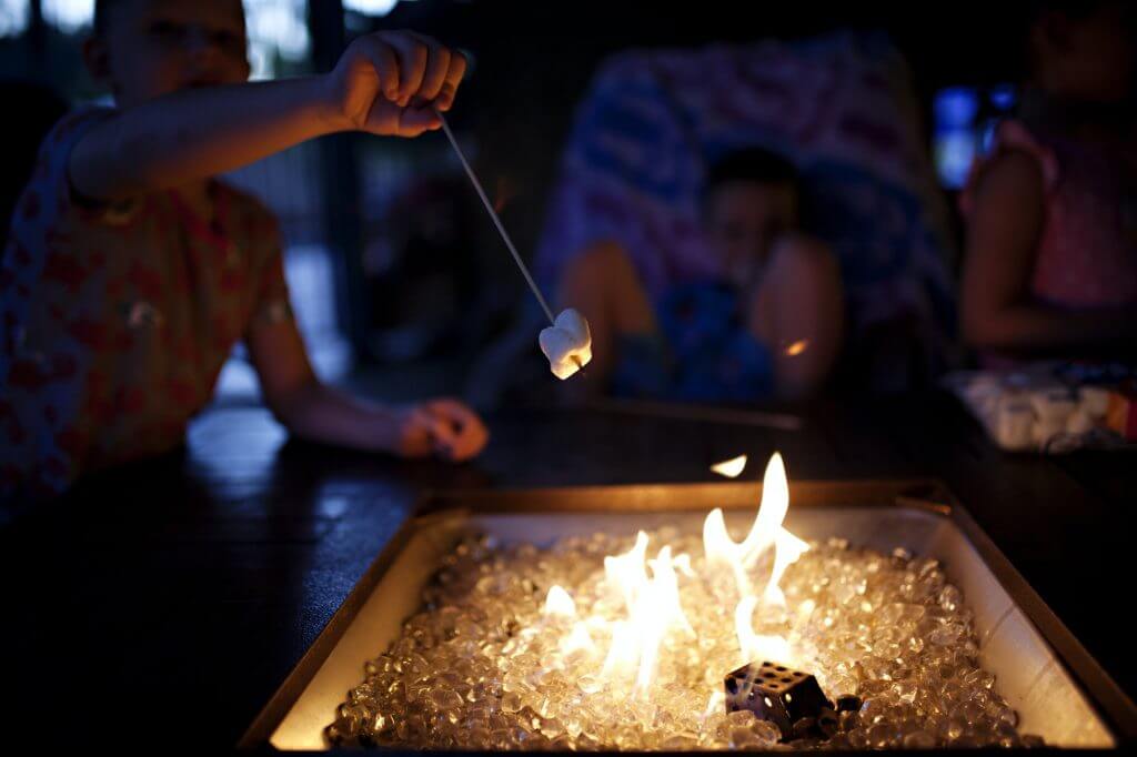 Kids sitting around a patio fire pit with flames roasting s’mores.